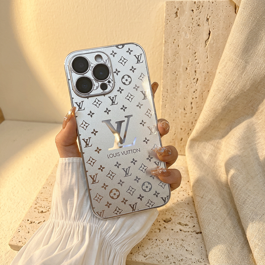 Silver Louis Vuitton monogram design iPhone case held upright by a hand with decorated nails and a white sleeve, showing dark floral motifs and a large reflective LV detail.