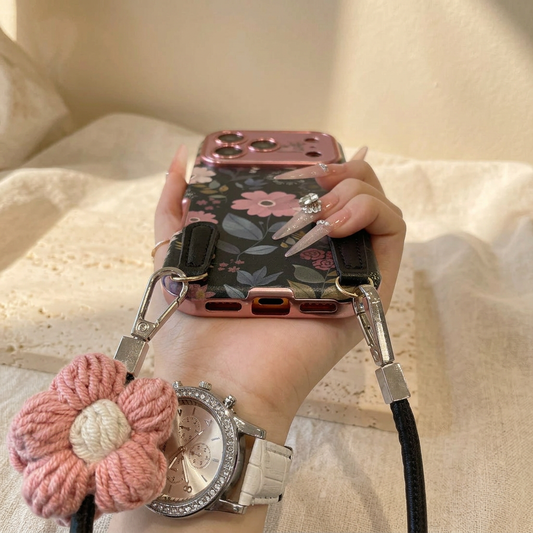 Close-up of the floral case bottom edge, charging-port opening, black attachment tabs, silver-coloured strap clips and pink crochet flower with a cream centre.