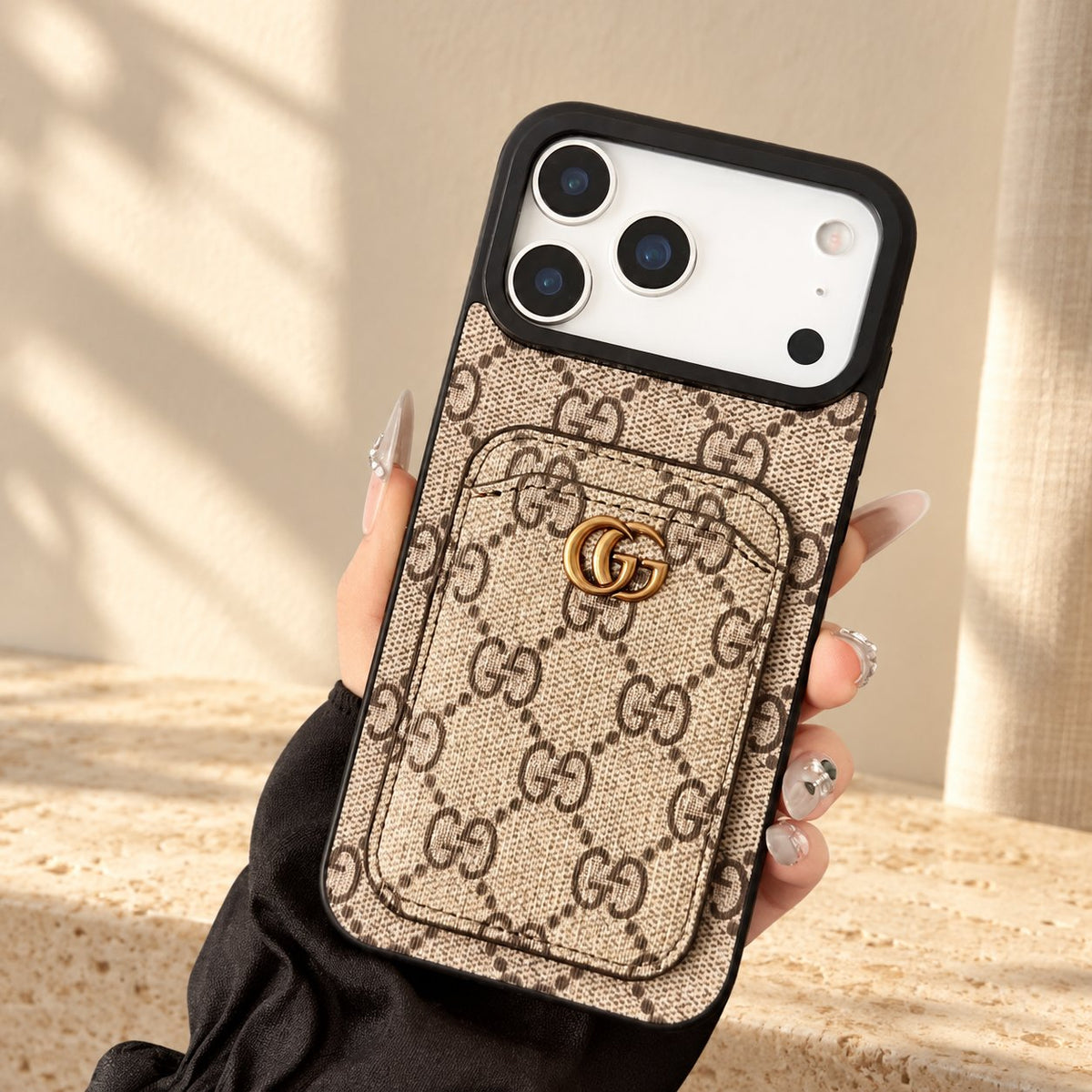 Hand holding the beige GG monogram design iPhone case with a stitched rear card pocket, gold-coloured GG emblem and display phone in a warm ivory setting.