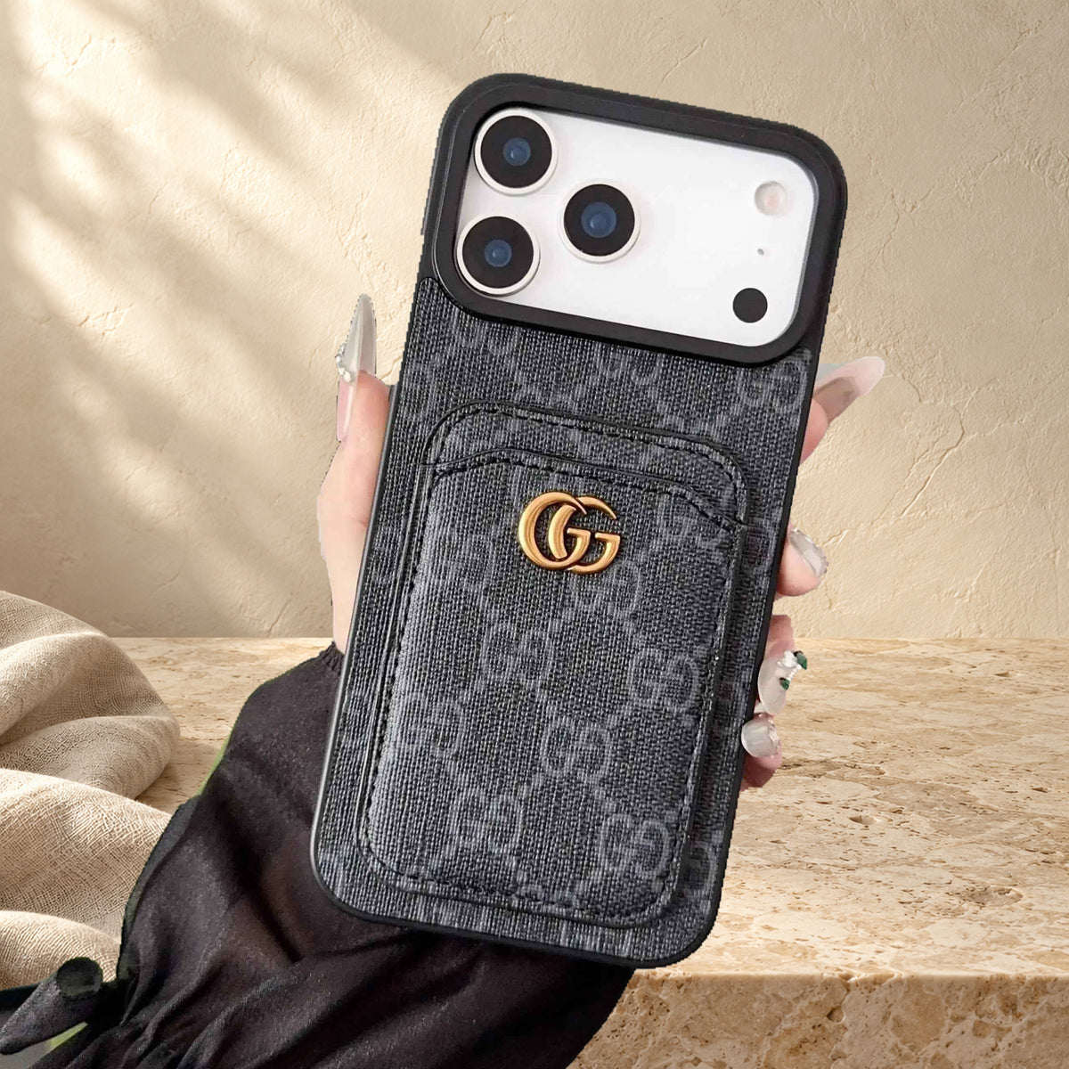Hand holding the dark GG monogram design case, showing the stitched rear card pocket, gold-coloured emblem and display phone.