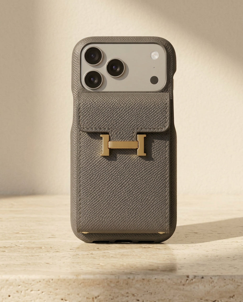 Grey grained iPhone case with rear flap card pocket and gold-coloured H buckle, standing on cream travertine.