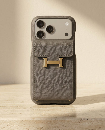 Grey grained iPhone case with rear flap card pocket and gold-coloured H buckle, standing on cream travertine.