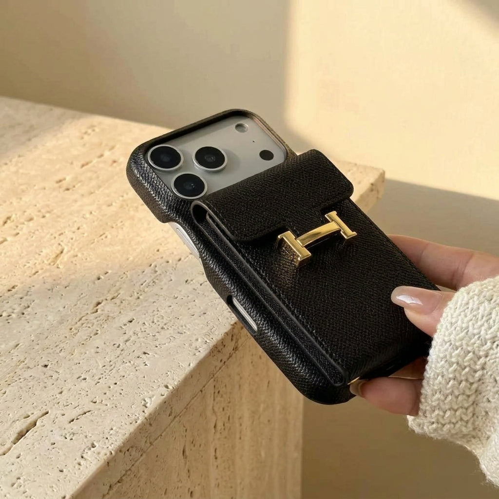 Black iPhone case with gold-coloured H buckle, held beside a cream travertine ledge.
