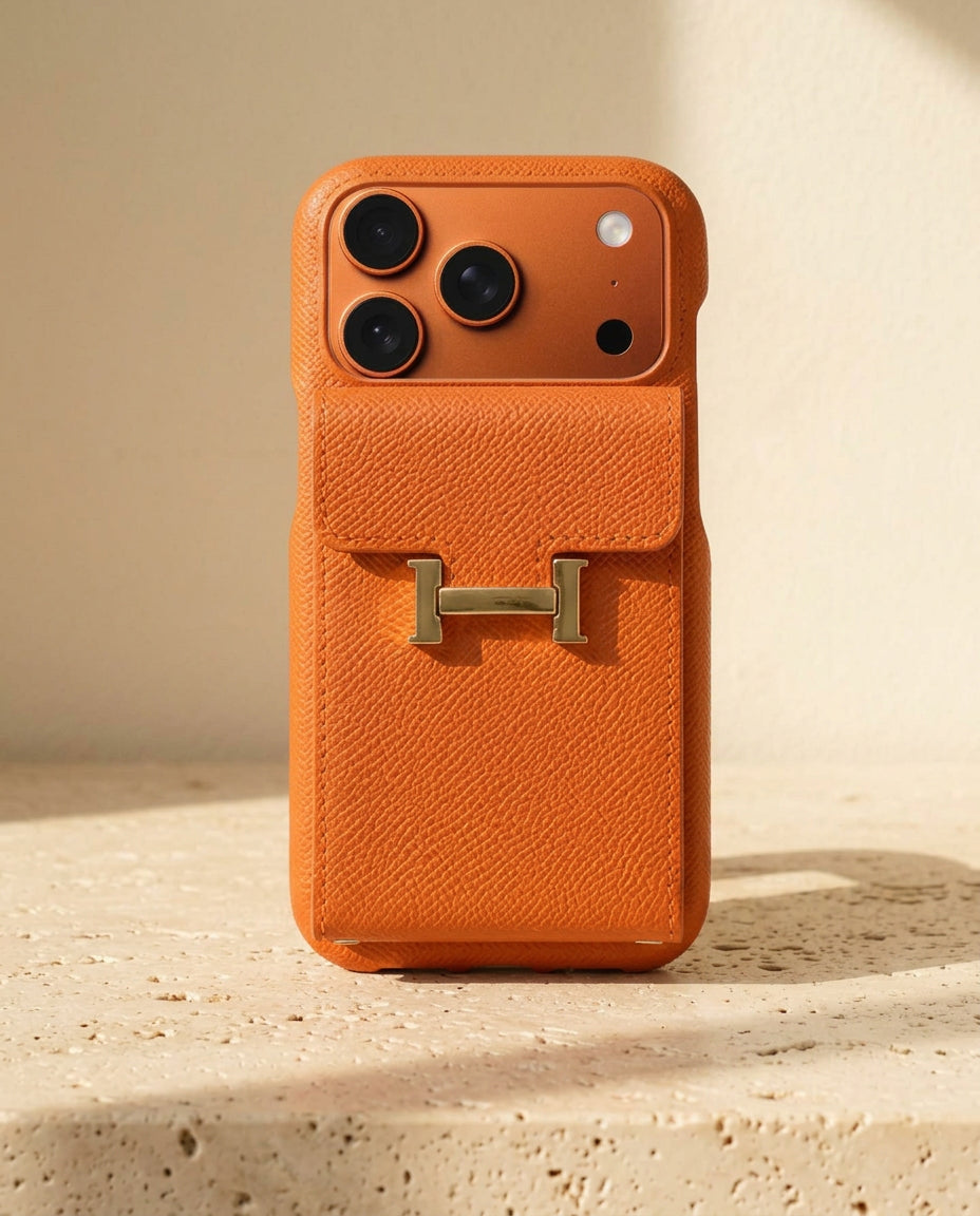 Orange grained iPhone case with rear flap card pocket and gold-coloured H buckle, standing on cream travertine.