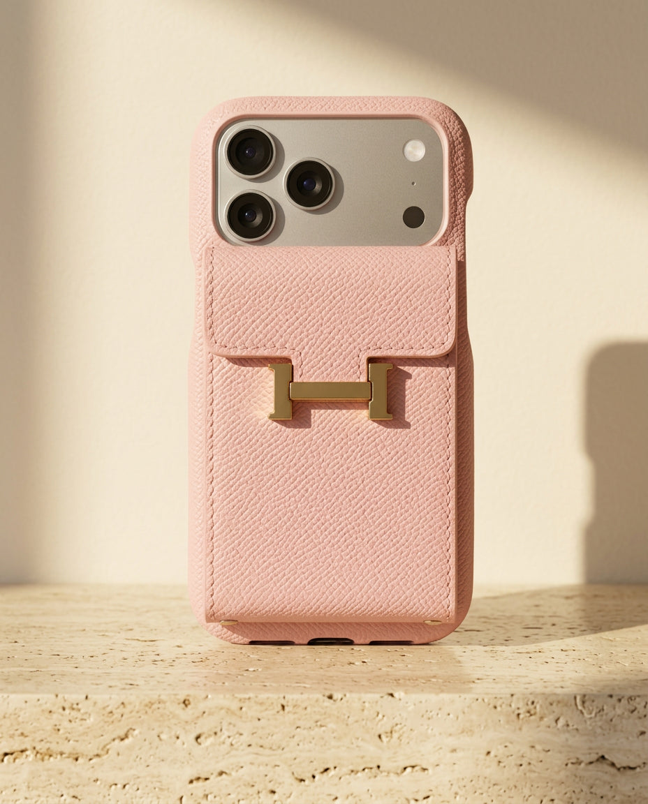 Pink grained iPhone case with rear flap card pocket and gold-coloured H buckle, standing on cream travertine.