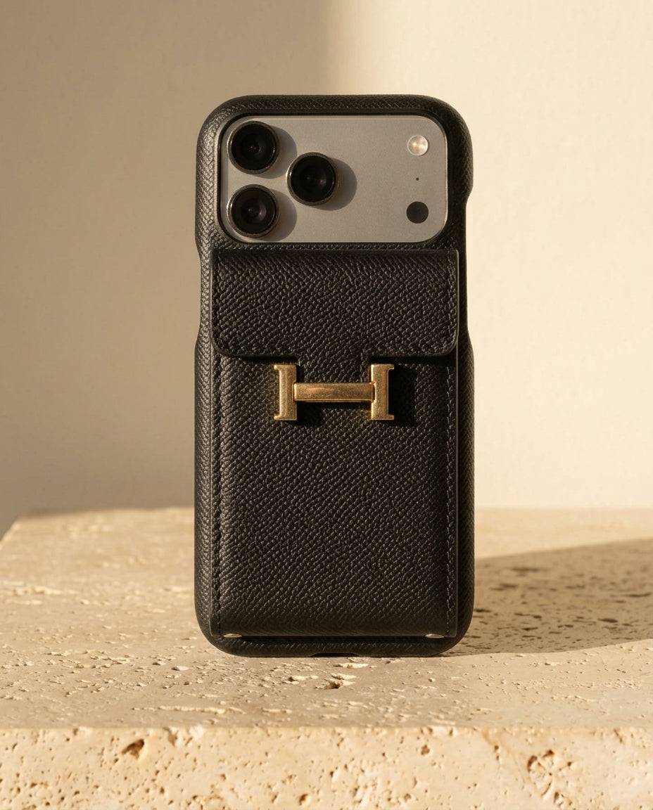Black grained iPhone case with rear flap card pocket and gold-coloured H buckle, standing on cream travertine.