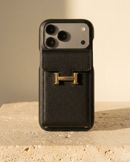 Black grained iPhone case with rear flap card pocket and gold-coloured H buckle, standing on cream travertine.
