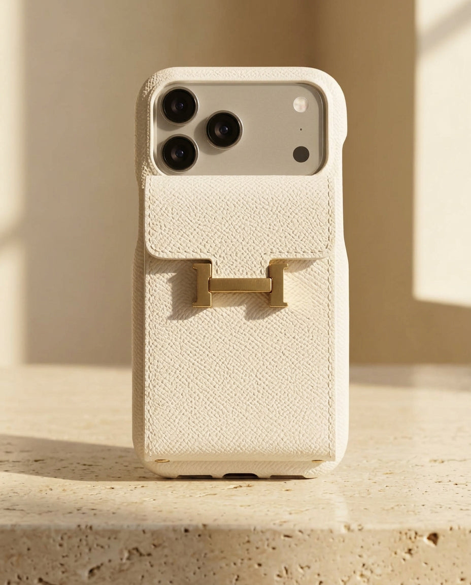 White grained iPhone case with rear flap card pocket and gold-coloured H buckle, standing on cream travertine.
