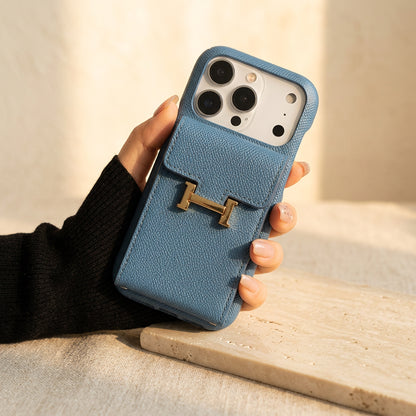 Blue grained iPhone case with rear flap card pocket and gold-coloured H buckle, held in hand over cream travertine.