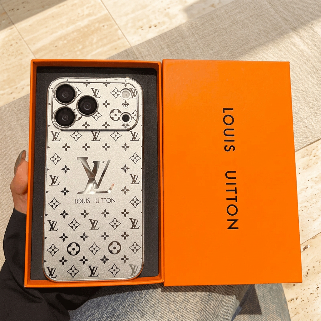 Silver Louis Vuitton monogram design iPhone case in an orange presentation tray beside an orange lid, showing contrasting dark motifs and a wide camera surround.