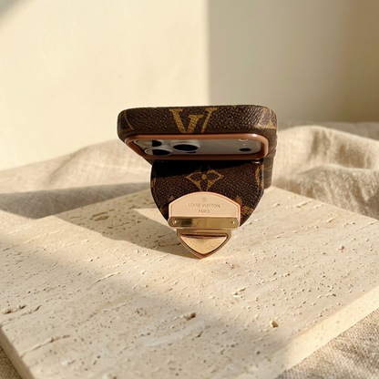 Low end-on view of the brown monogram case showing its upper edge, camera panel and projecting rear-pocket flap with gold-tone clasp.