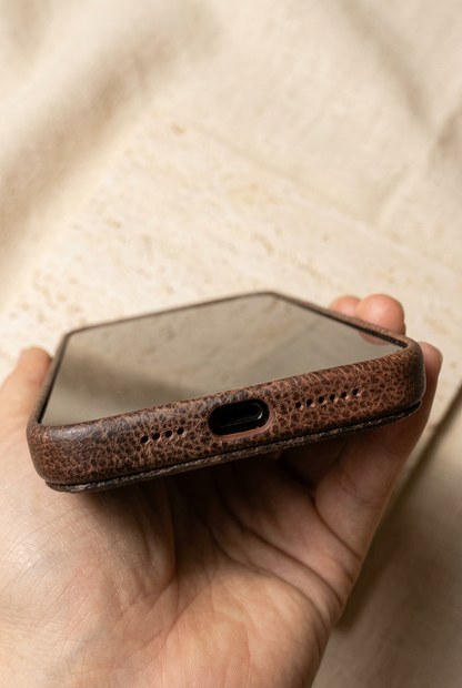 Hand holding a dark brown stitched case in a close-up of the screen, bottom charging opening and small speaker holes.