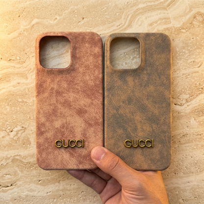 Hand holding peach-pink and khaki-brown distressed-look Gucci design cases side by side, showing gold-tone names and empty camera openings.
