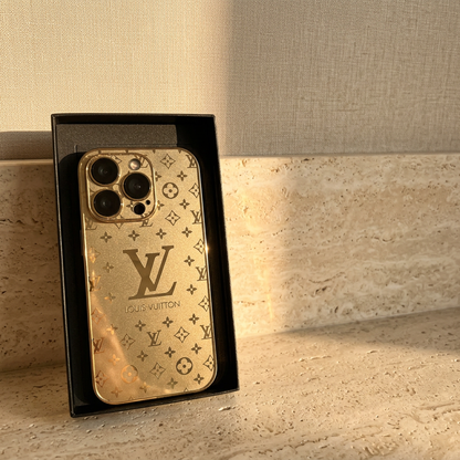 Gold Louis Vuitton monogram design iPhone case upright in a black display tray, showing a large central LV motif and compact patterned camera surround.