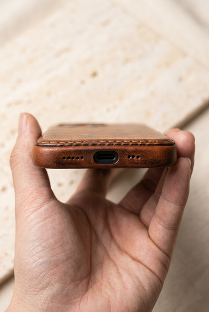 Close-up of a hand holding the brown case with rear-panel stitching above the bottom charging opening and speaker holes.