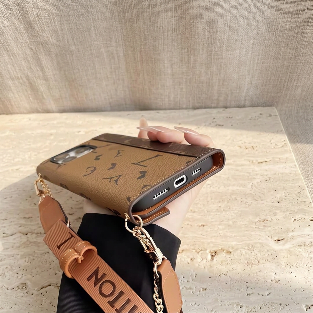 Hand-held bottom view of the brown monogram folio showing the phone's charging port, speaker openings, strap clips and chain.