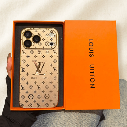 Gold Louis Vuitton monogram design iPhone case in an orange presentation tray beside its orange lid, showing a large LV motif and wide patterned camera panel.