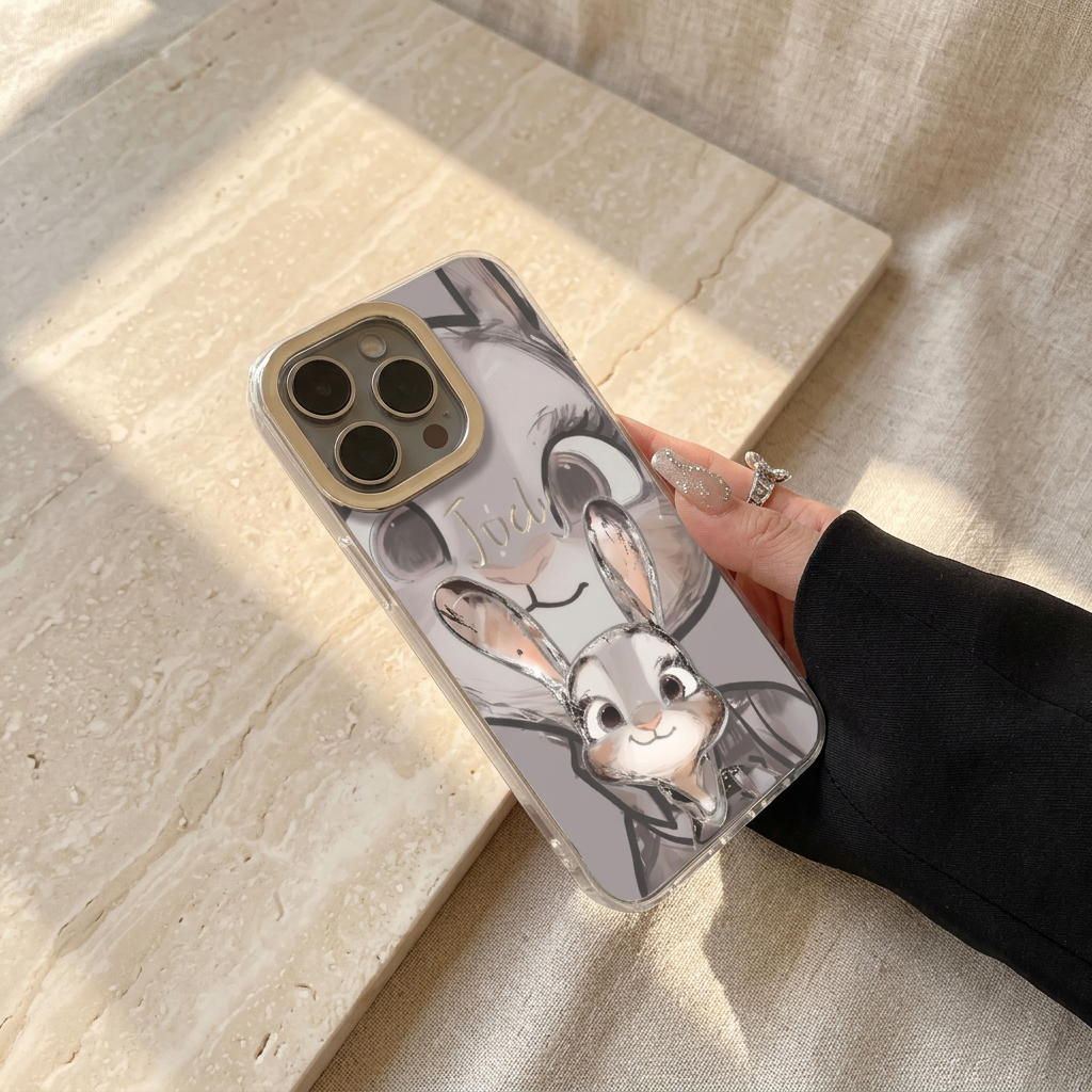 Angled hand-held Judy case with grey rabbit portraits, a silver-tone Judy name and clear-looking edging, held beside a glitter nail and butterfly ring.