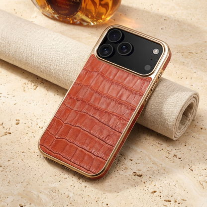 Close diagonal rear view of the reddish-brown croc-pattern case resting across rolled linen, with gold-tone camera trim.